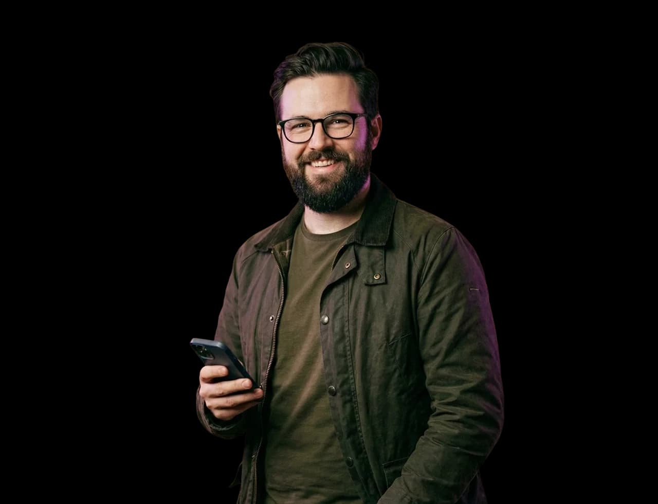 Fiatal szemüveges férfi mobiltelefonnal a kezében mosolyog.